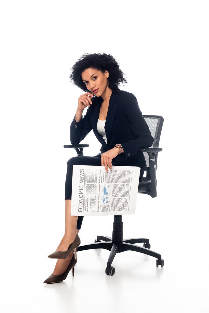 Африканская бизнесвумен в офисном кресле держит газету и смотрит в камеру на белом  - Фото, изображение
