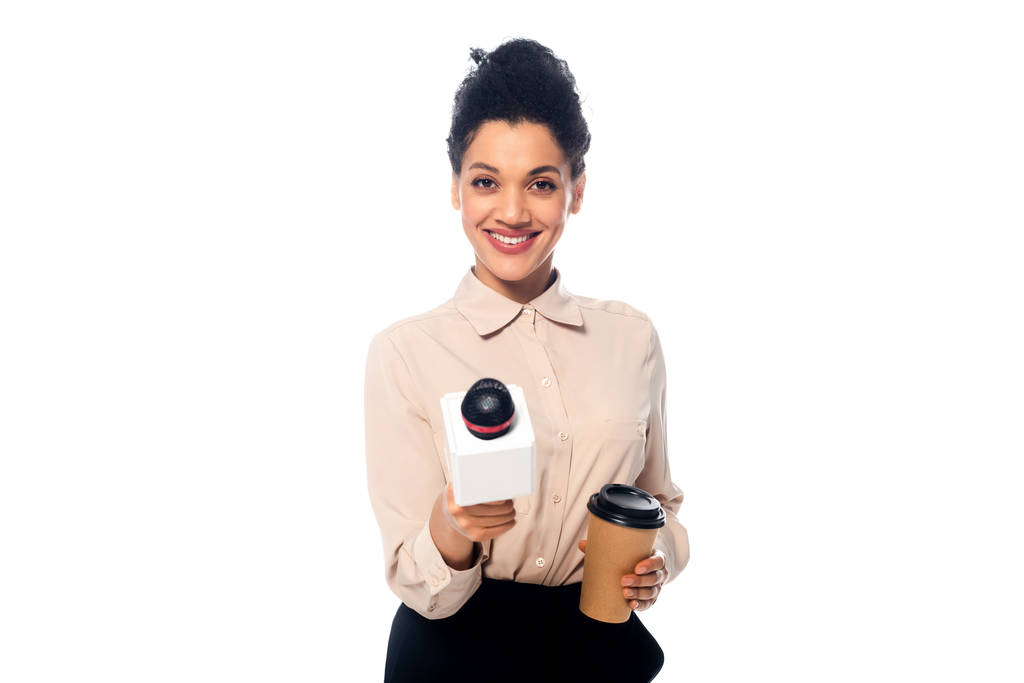 Vista frontal de periodista afroamericano con taza de café desechable sonriendo y mostrando micrófono aislado en blanco
 - Foto, Imagen
