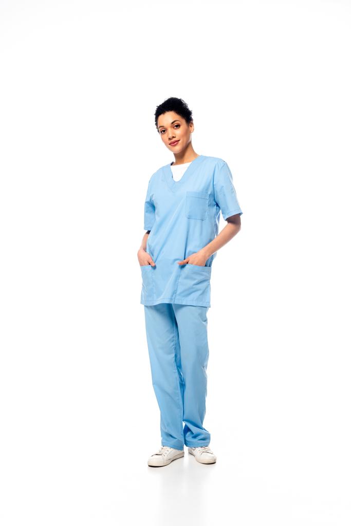 Vista completa de la enfermera afroamericana con las manos en el bolsillo mirando a la cámara sobre fondo blanco
 - Foto, imagen