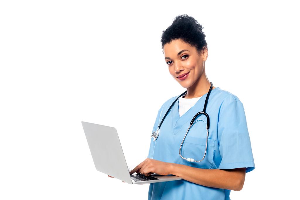 Afro-Amerikaanse verpleegster met stethoscoop en laptop glimlachen en kijken naar camera geïsoleerd op wit - Foto, afbeelding