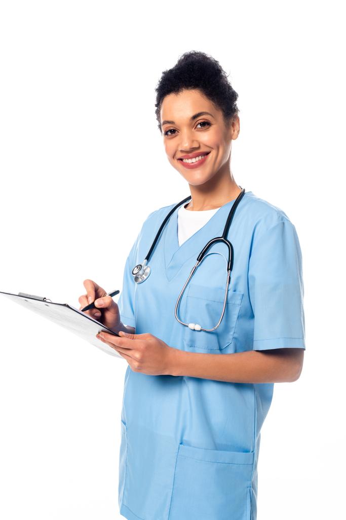 Afro-Amerikaanse verpleegster met stethoscoop schrijven op klembord en glimlachen geïsoleerd op wit - Foto, afbeelding