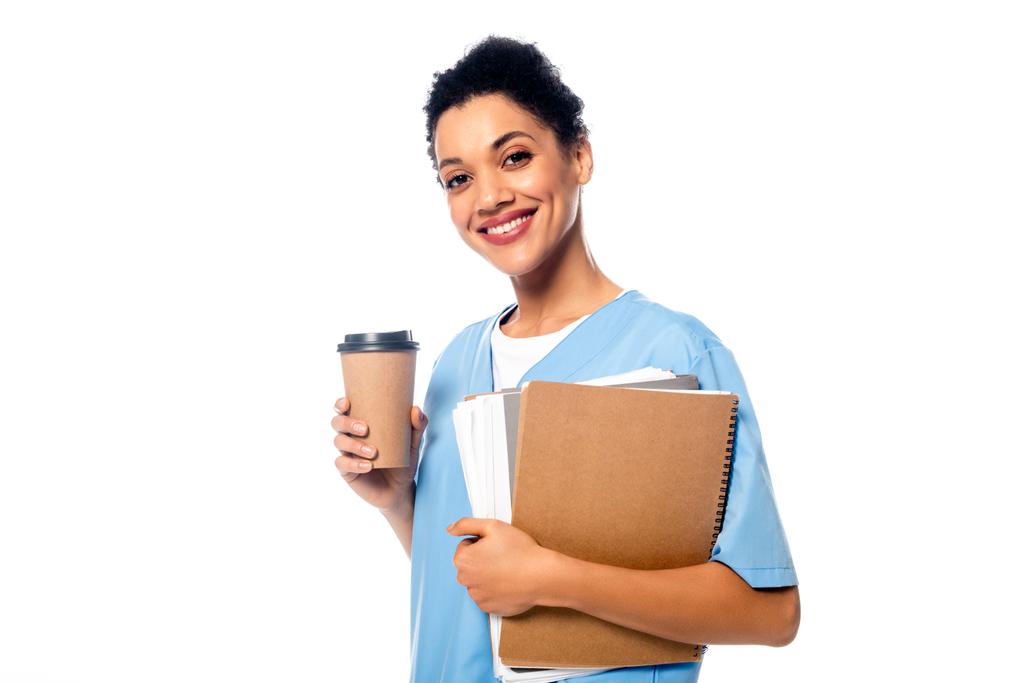 Szczęśliwa i pozytywna afrykańska pielęgniarka z papierami i jednorazową filiżanką kawy patrzącą na kamerę odizolowaną na biało - Zdjęcie, obraz