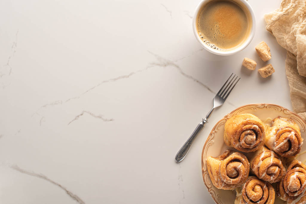 vista superior de rollos de canela caseros frescos en la superficie de mármol con taza de café, tenedor y tela
 - Foto, imagen