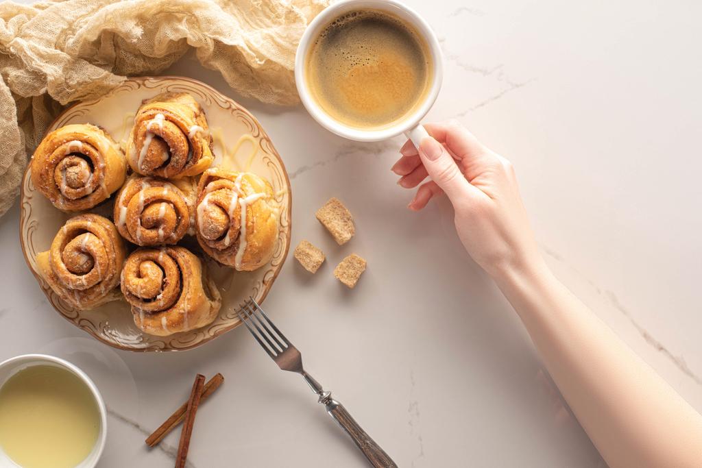 vista recortada de la mujer sosteniendo taza de café cerca de rollos de canela caseros en la superficie de mármol con tenedor, leche condensada y tela
 - Foto, imagen