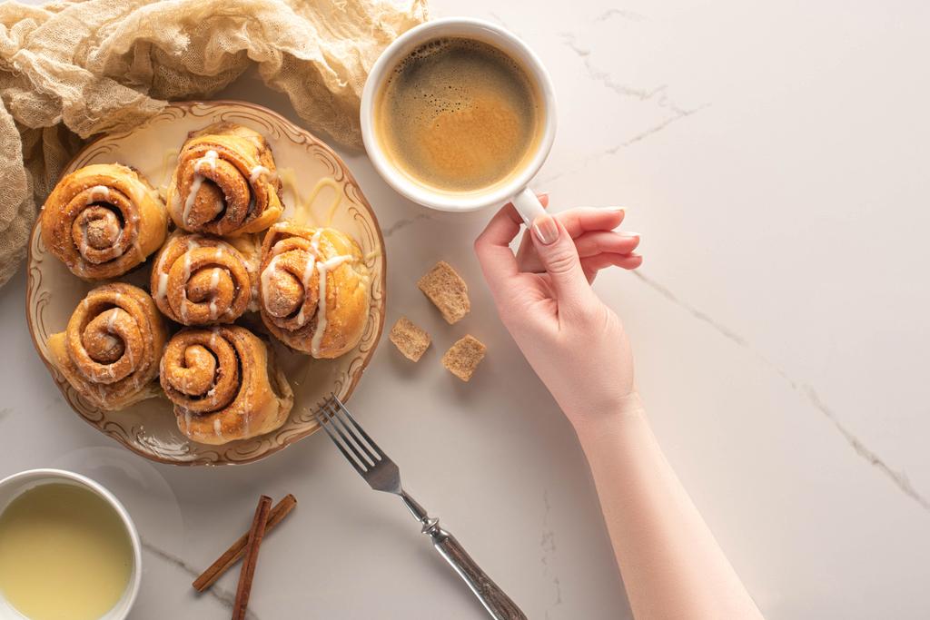 cropped view of woman holding cup of coffee near homemade cinnamon rolls on marble surface with fork, condensed milk and cloth - Photo, Image