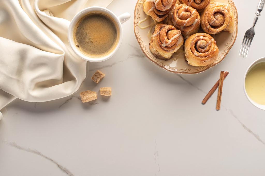 top view of fresh homemade cinnamon rolls on marble surface with cup of coffee, brown sugar, condensed milk, fork and silk cloth - Photo, Image