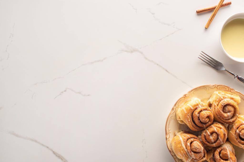 πάνω όψη φρέσκων χειροποίητων ρολών κανέλας σε μαρμάρινη επιφάνεια με συμπυκνωμένο γάλα, πιρούνι - Φωτογραφία, εικόνα