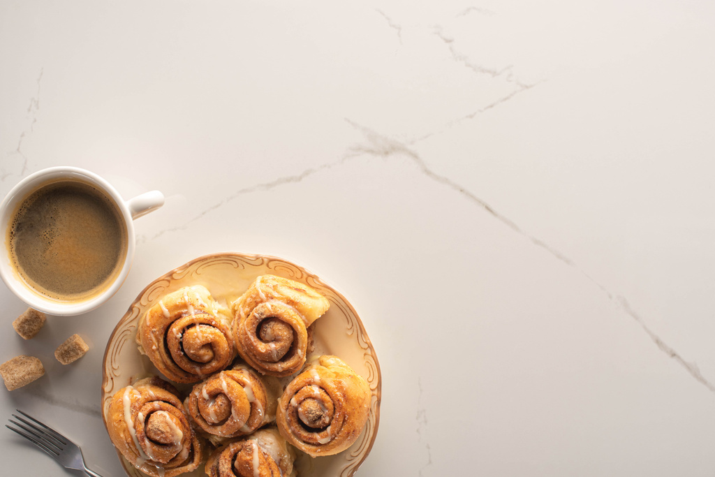 Draufsicht auf frische hausgemachte Zimtrollen auf Marmoroberfläche mit Tasse Kaffee, Gabel und braunem Zucker - Foto, Bild