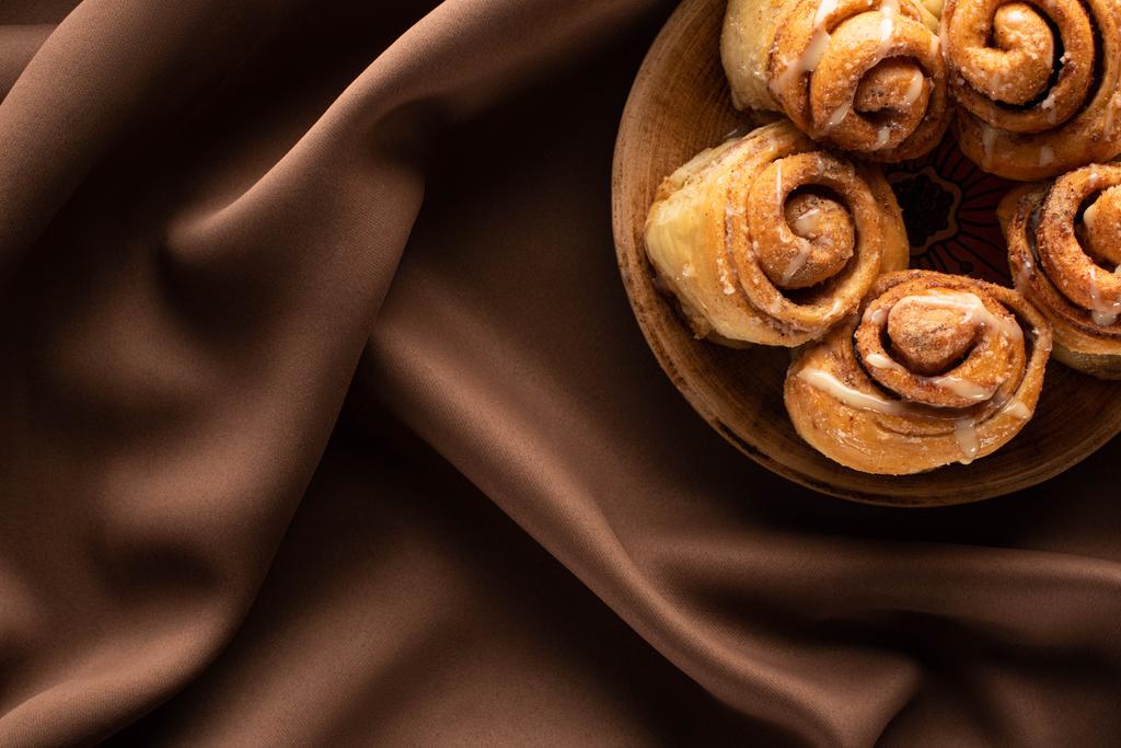 top view of fresh homemade cinnamon rolls on plate on silk brown cloth - Photo, Image