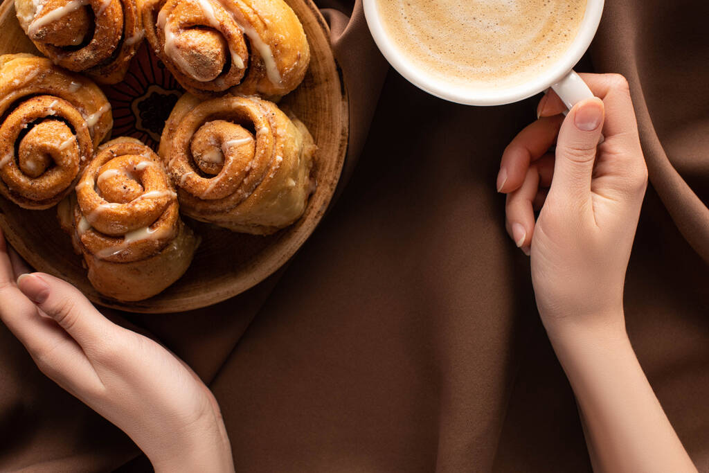 cropped view of woman holding plate with fresh homemade cinnamon rolls near coffee on silk brown cloth - Photo, Image