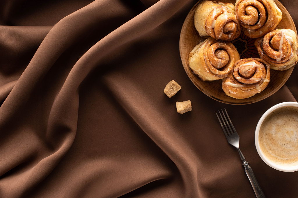 top view of fresh homemade cinnamon rolls on silk brown cloth with brown sugar, fork and coffee - Photo, Image