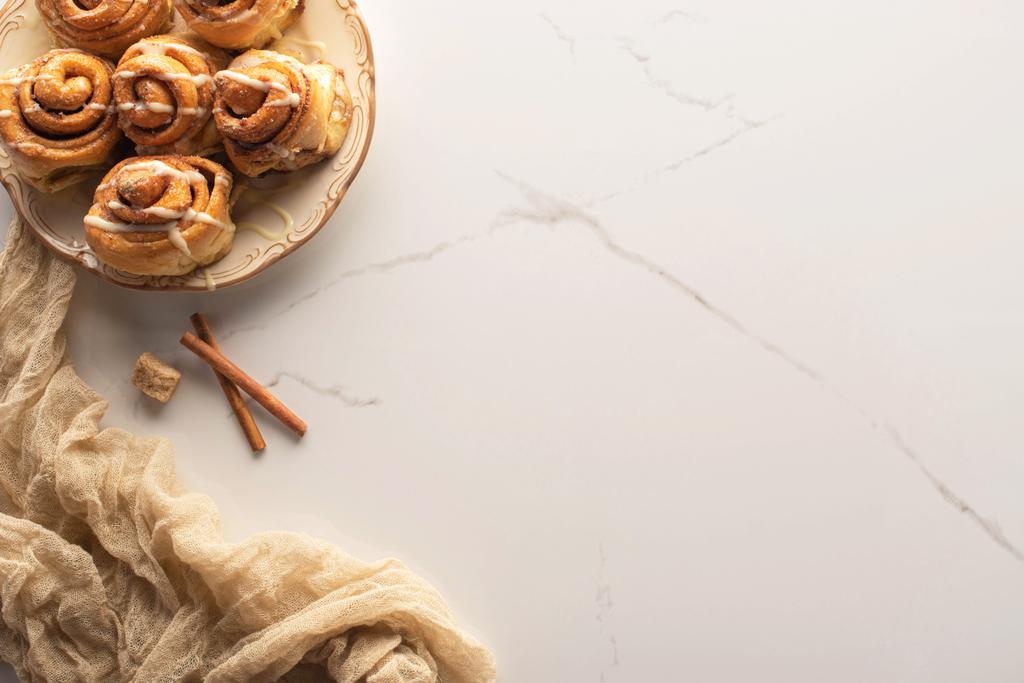 πάνω όψη φρέσκων χειροποίητων ρολών κανέλας σε μαρμάρινη επιφάνεια με καστανή ζάχαρη, ξυλάκια κανέλας και ύφασμα - Φωτογραφία, εικόνα