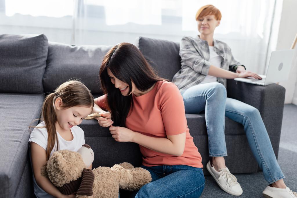 Szelektív fókusz mosolygós anya közelében lánya játékmackó és szülő laptop kanapén otthon - Fotó, kép