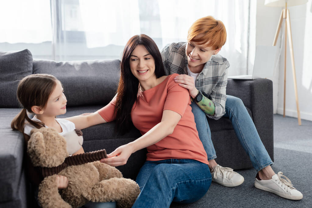 Sonriendo familia del mismo sexo sentado cerca de la hija con oso de peluche en la sala de estar
 - Foto, Imagen