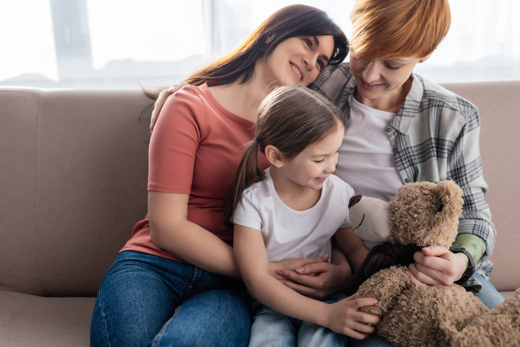 Smiling same sex parents sitting near daughter with teddy bear on couch - Photo, Image