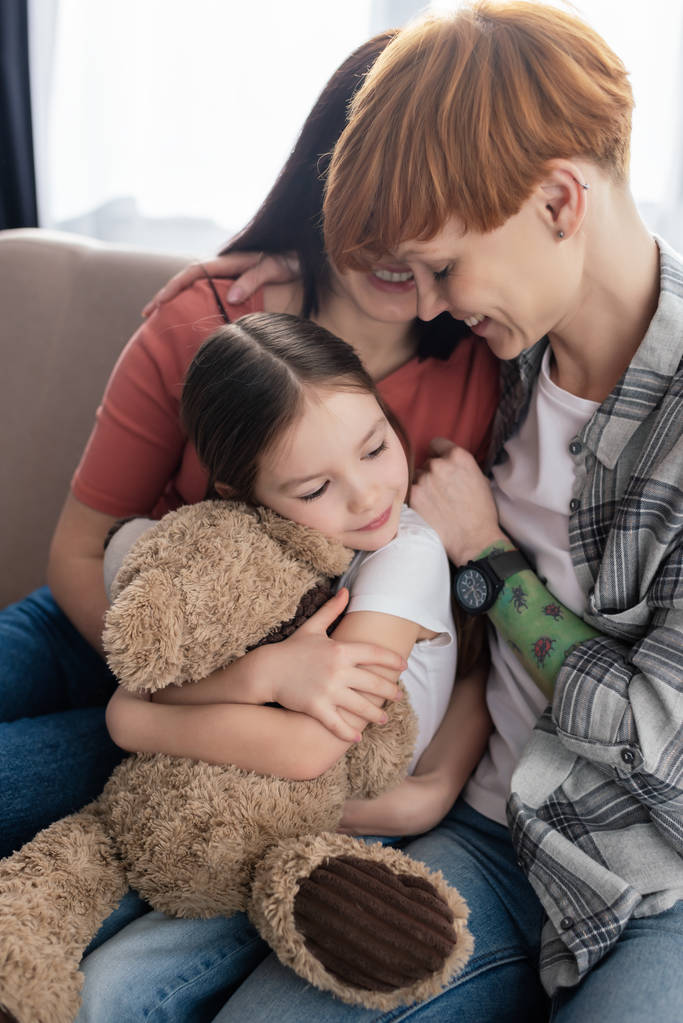 Feliz mismo sexo padres abrazando hija con osito de peluche en sofá
 - Foto, imagen