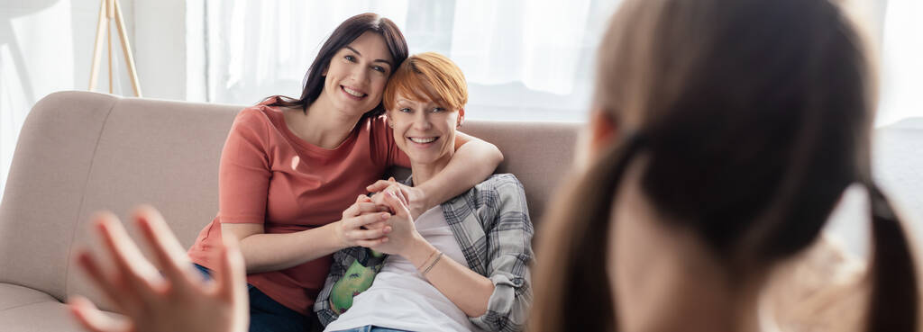 Выборочный фокус улыбающихся матерей, смотрящих на дочь, машущую рукой дома, панорамный снимок
 - Фото, изображение