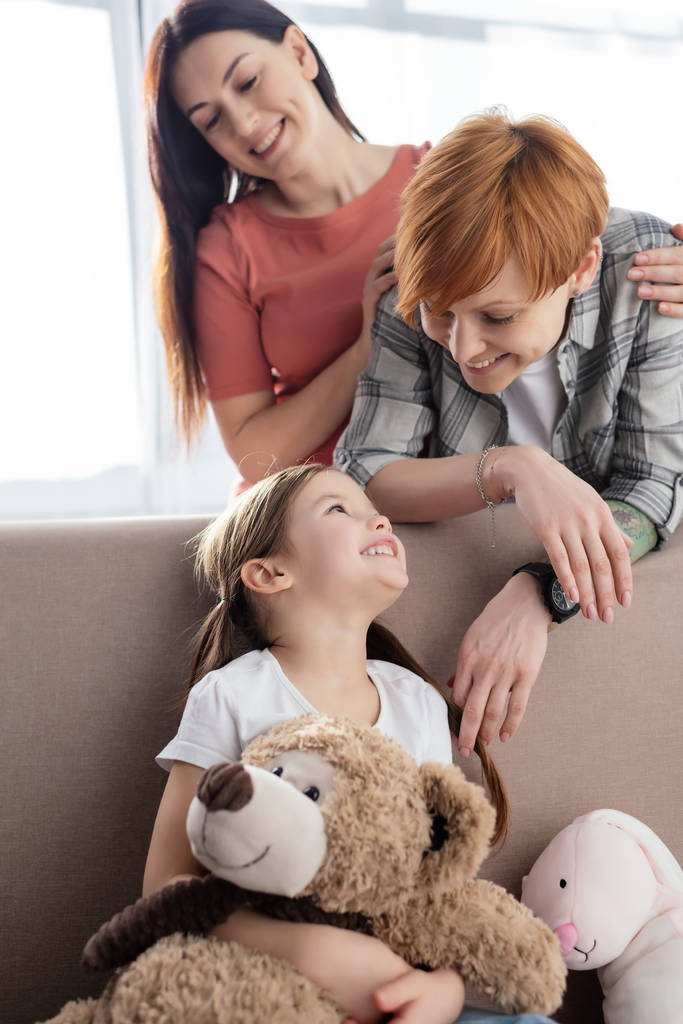 Selektywne skupienie wesołego dziecka z miękkimi zabawkami na kanapie patrząc na rodziców tej samej płci w salonie - Zdjęcie, obraz