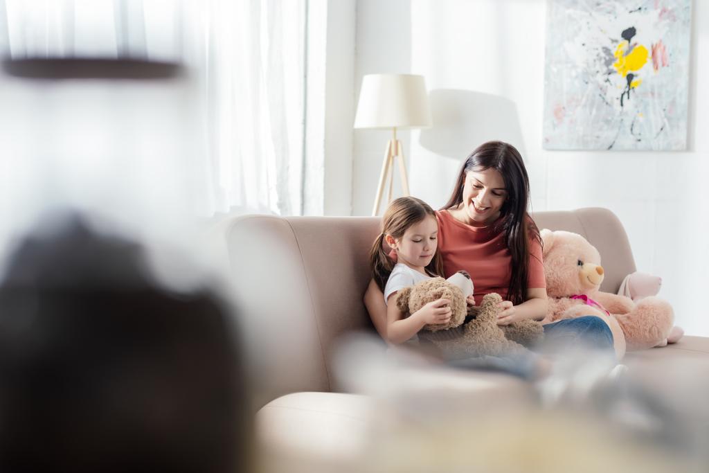 Selectieve focus van kind spelen met teddybeer in de buurt van gelukkige moeder op de bank in de woonkamer - Foto, afbeelding