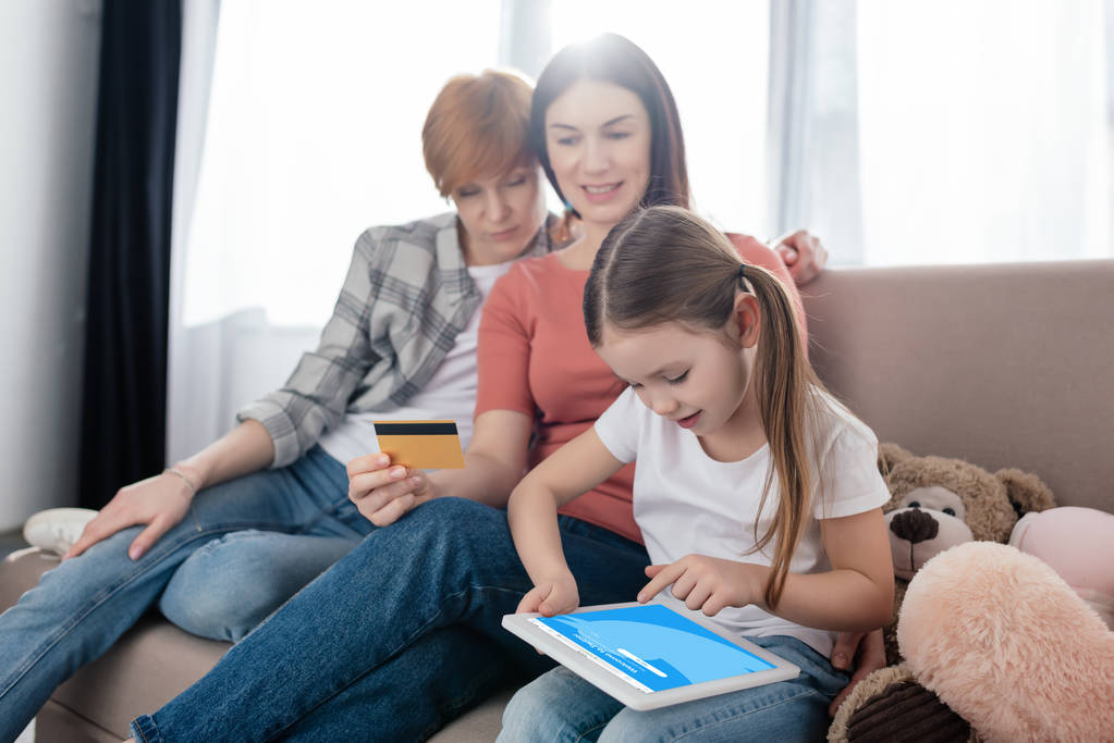 KYIV, UKRAINE - FEBRUARY 10, 2020: Child using digital tablet with twitter app on screen near smiling same sex parents with credit card on sofa - Fotó, kép