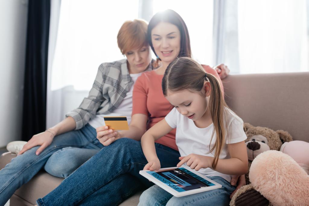 Kind nutzt digitales Tablet mit Buchungs-App am Bildschirm in der Nähe von Müttern mit Kreditkarte zu Hause - Foto, Bild