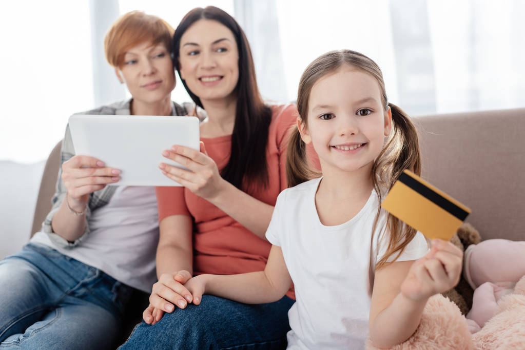 Foco seletivo de criança segurando cartão de crédito e sorrindo para a câmera perto de mães com tablet digital no sofá
  - Foto, Imagem