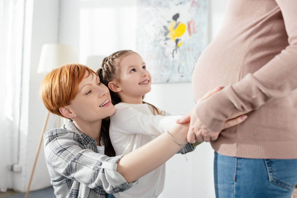 Vue latérale de la mère et de la fille souriantes touchant le ventre de la femme enceinte à la maison
  - Photo, image