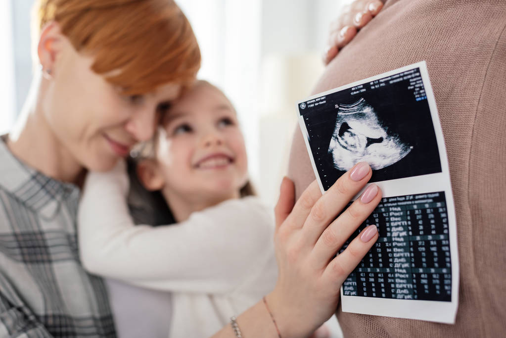 Selective focus of mother hugging smiling daughter and holding ultrasound scan of baby near belly of pregnant woman  - Photo, Image