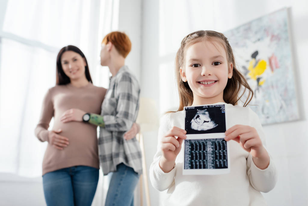 Valikoiva painopiste hymyilevä lapsi tilalla ultraääni skannaus vauva lähellä nainen käsittää raskaana tyttöystävä
  - Valokuva, kuva