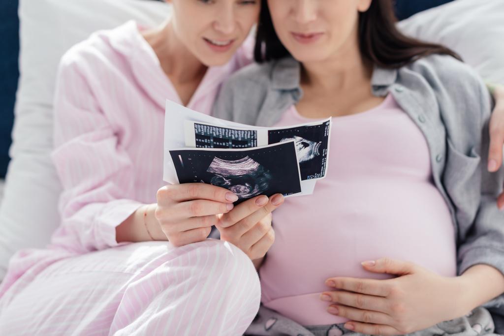 Selective focus of smiling same sex couple holding ultrasound scans of baby on bed isolated on blue  - Photo, Image