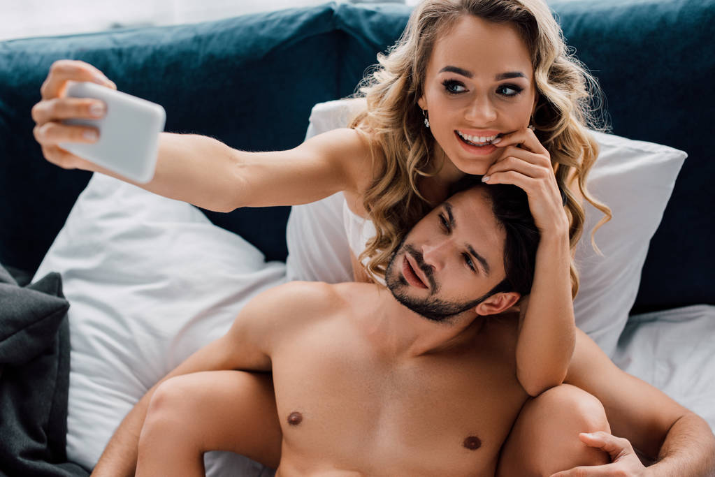 Mulher sorridente bonita tomando selfie com smartphone perto do namorado muscular na cama
  - Foto, Imagem