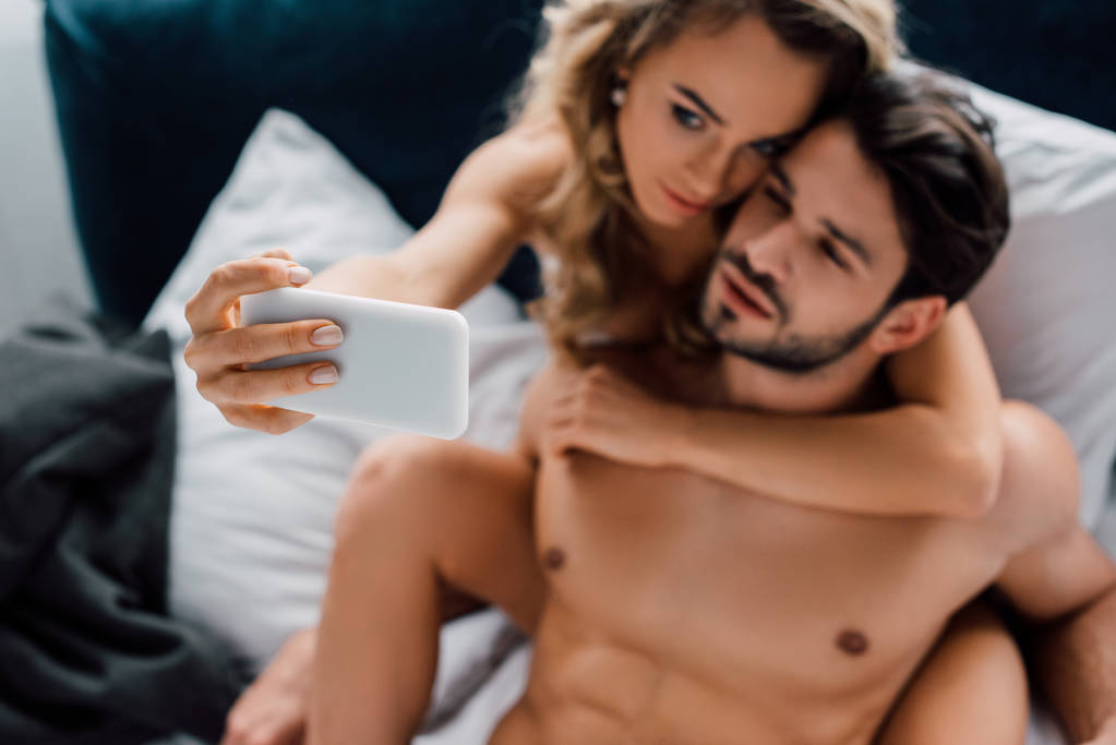Güzel bir kadının kaslı erkek arkadaşına sarıldığı seçmeli bir odak noktası. Gri yatakta akıllı telefonuyla selfie çekerken. - Fotoğraf, Görsel