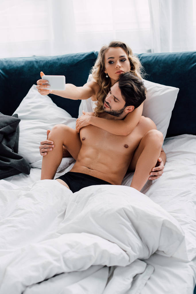 Houkutteleva nainen ottaa selfie älypuhelimen lähellä lihaksikas poikaystävä sängyssä
  - Valokuva, kuva