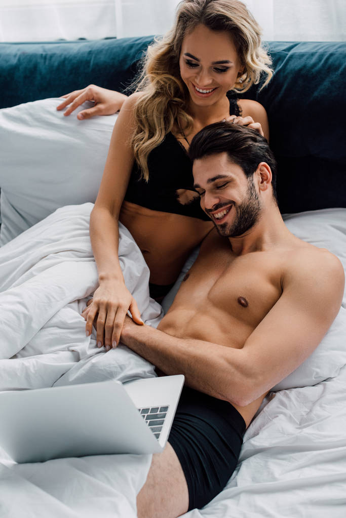 glimlachende vrouw in beha kijken naar laptop in de buurt van knappe vriendje op bed  - Foto, afbeelding