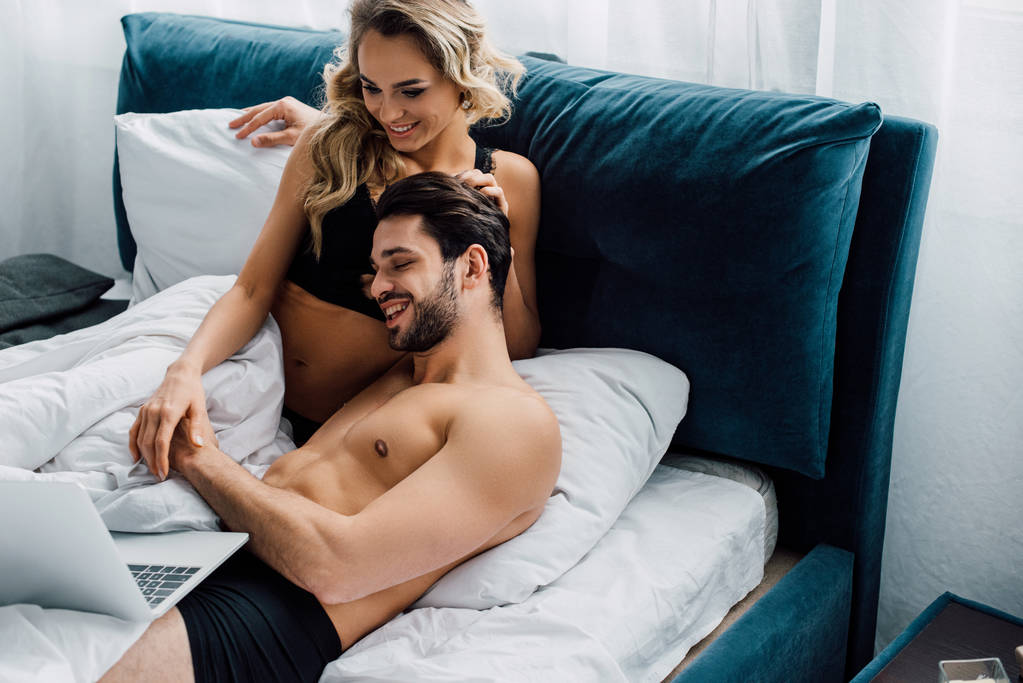 Sexy mulher tocando sorrindo namorado enquanto olha para laptop na cama
  - Foto, Imagem