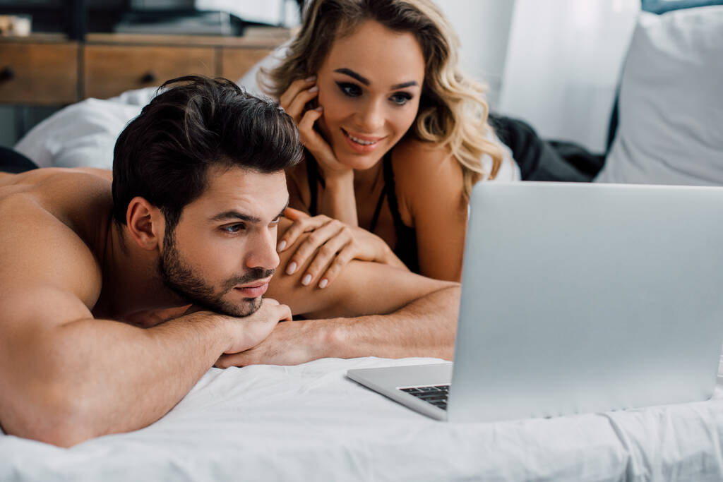 Knappe man kijkt naar laptop in de buurt glimlachende vrouw op bed  - Foto, afbeelding