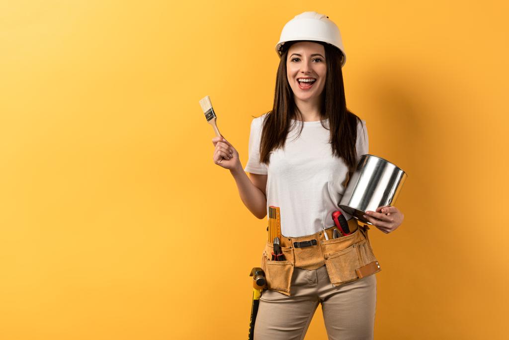 usmívající se údržbářka drží štětec a plechovku barvy na žlutém pozadí  - Fotografie, Obrázek