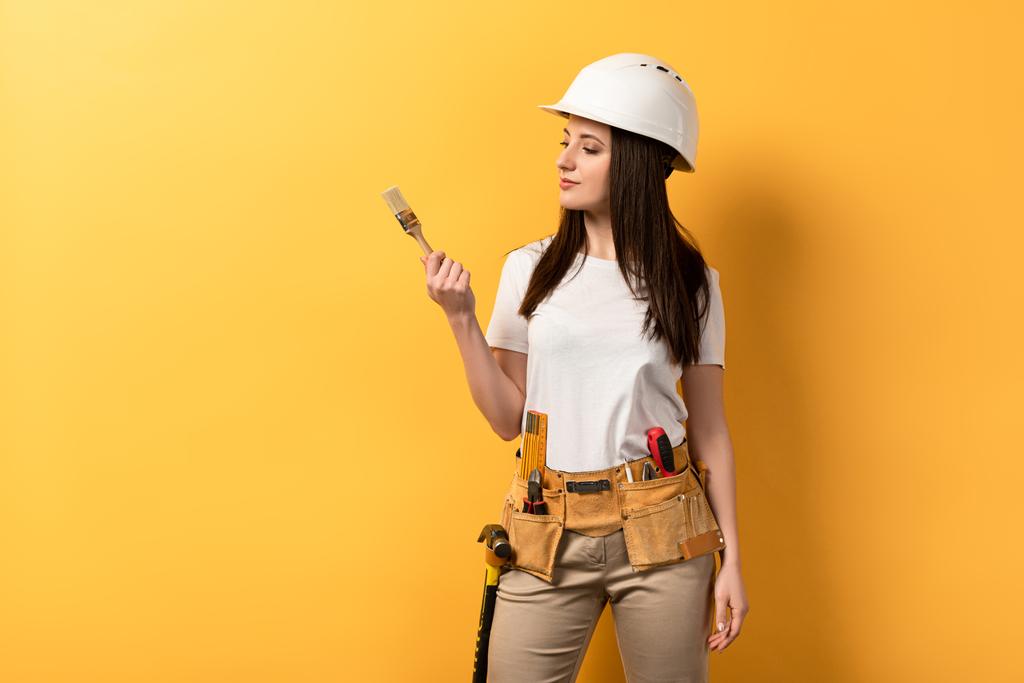 серьезная работница держит и смотрит на кисть краски на желтом фоне
  - Фото, изображение