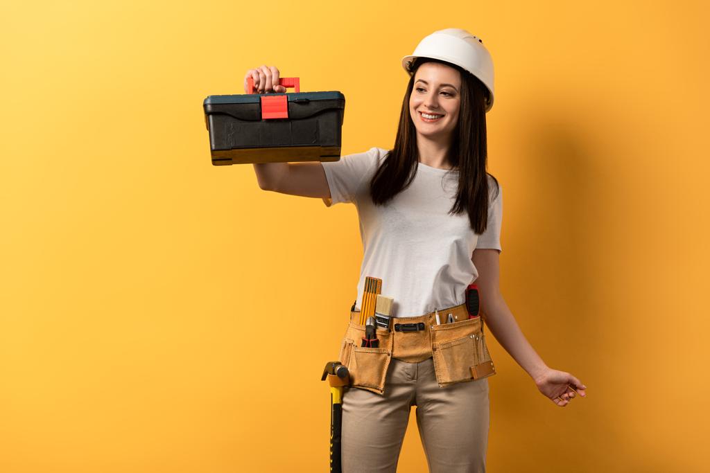 sonriente manitas en casco sosteniendo caja de herramientas sobre fondo amarillo
  - Foto, Imagen