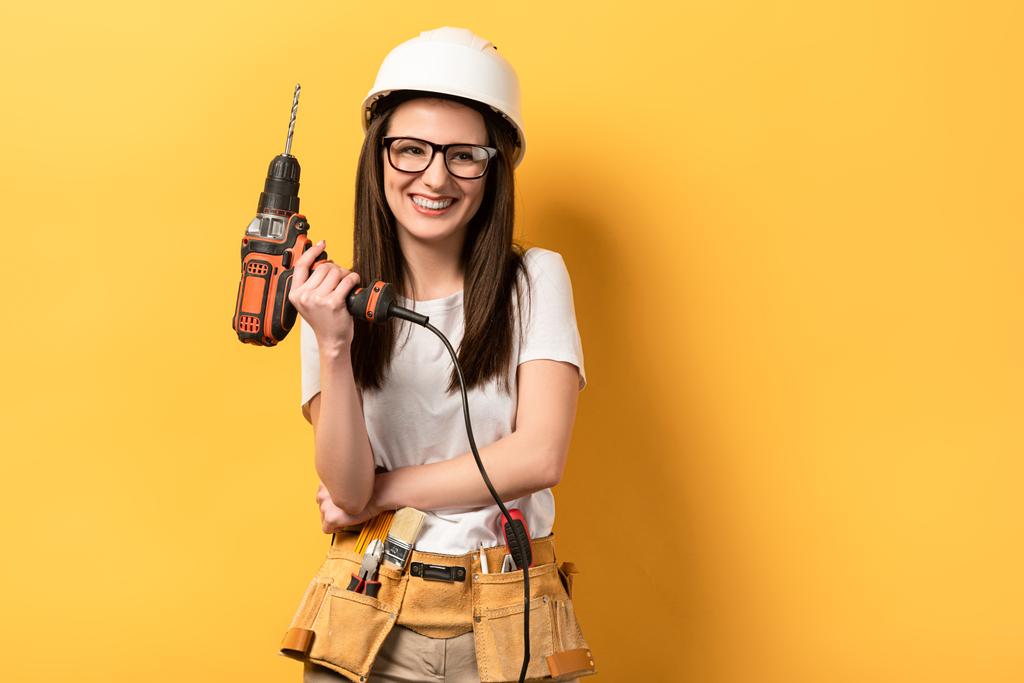 smiling handywoman in helmet holding drill on yellow background  - Photo, Image