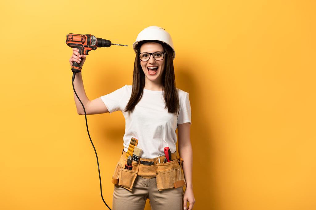 lächelnde Handarbeiterin mit Bohrmaschine und Blick in die Kamera auf gelbem Hintergrund  - Foto, Bild