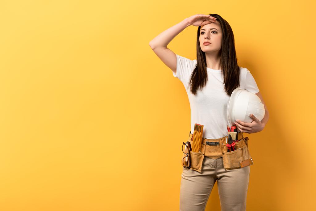 vážná údržbářka drží helmu a dívá se jinam na žluté pozadí  - Fotografie, Obrázek