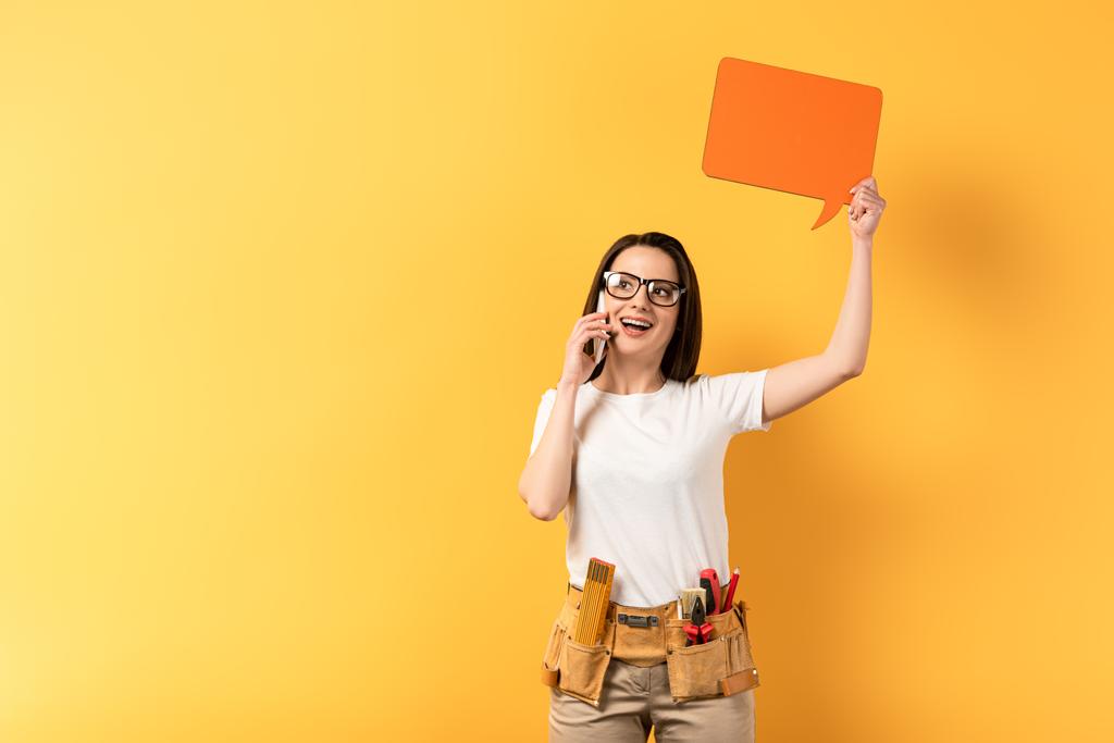 Gülümseyen tamirci kadın konuşma baloncuğu tutuyor ve sarı arka planda akıllı telefondan konuşuyor  - Fotoğraf, Görsel