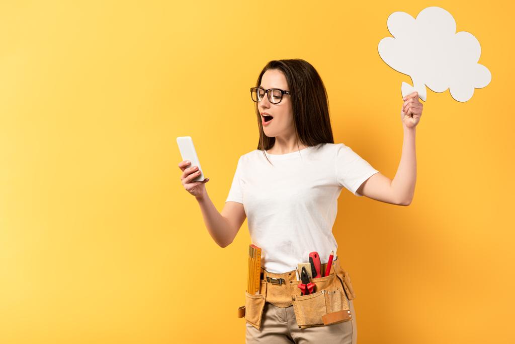 shocked repairwoman holding thought bubble and using smartphone on yellow background  - Photo, Image