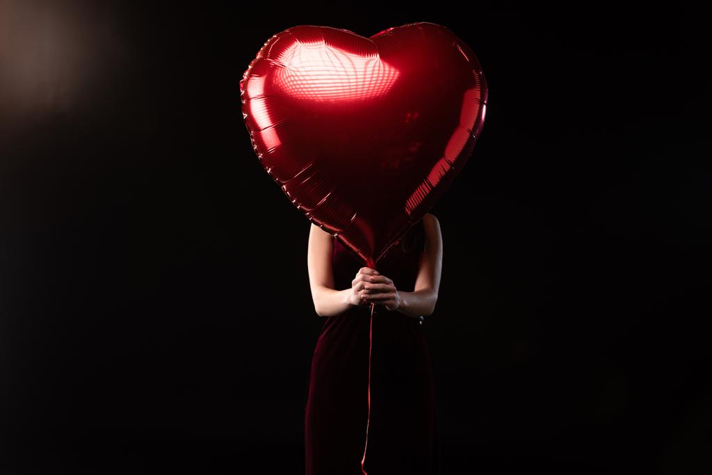 γυναίκα σε φόρεμα σκιάζει το πρόσωπο με μπαλόνι σε σχήμα καρδιάς σε 14 Φεβρουαρίου σε μαύρο φόντο  - Φωτογραφία, εικόνα