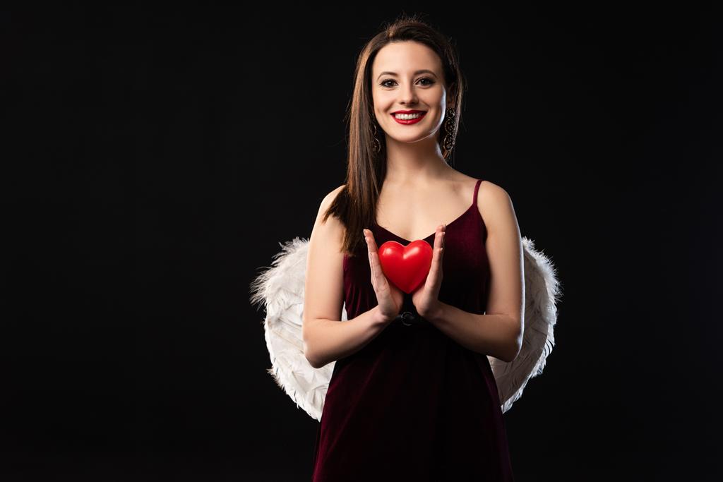 усміхнена жінка в одязі з крилами, що тримає модель у формі серця в 14 лютого, ізольована на чорному
 - Фото, зображення