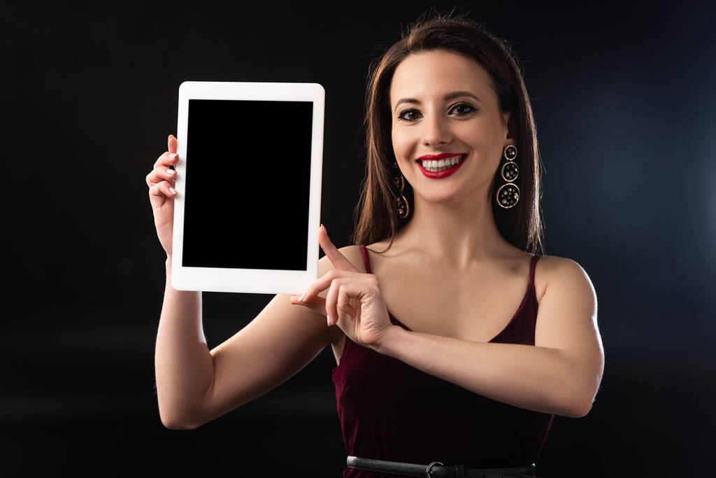 glimlachende vrouw in jurk houden digitale tablet op zwarte achtergrond  - Foto, afbeelding