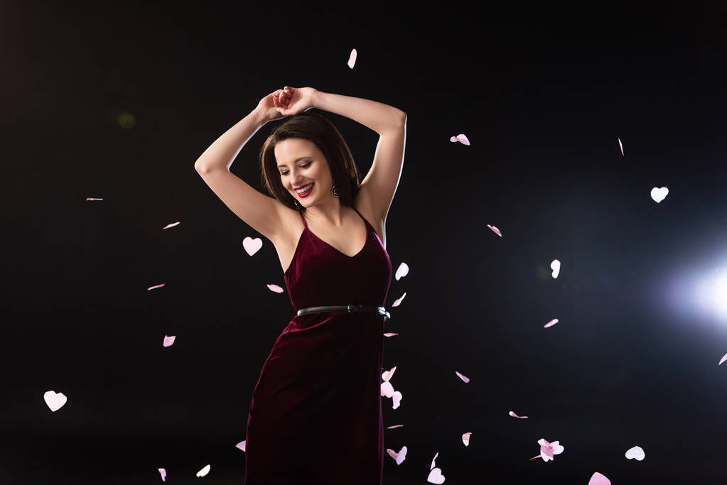 glimlachende vrouw in jurk staan in de buurt vallen confetti op zwarte achtergrond  - Foto, afbeelding