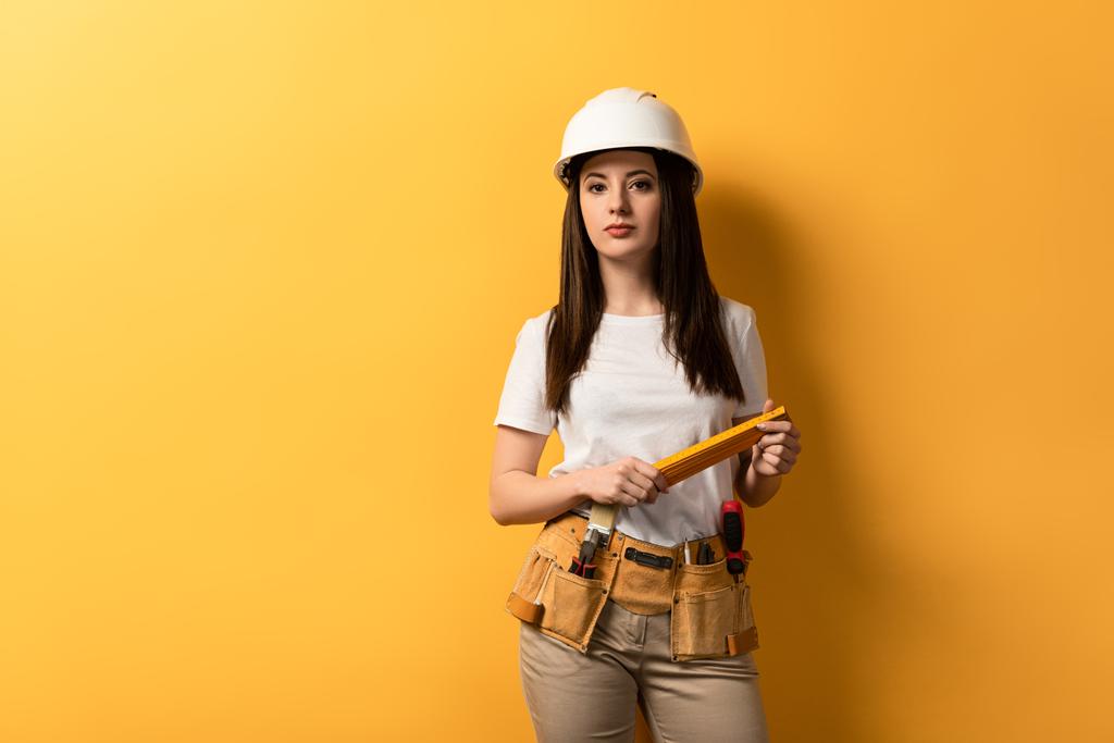 handywoman in helmet holding meter and looking at camera on yellow background  - Photo, Image