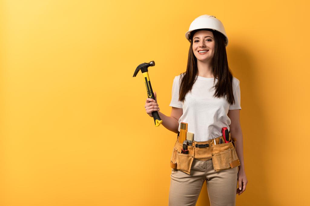 glimlachende handywoman in helm houden hamer op gele achtergrond  - Foto, afbeelding